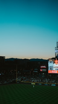 A baseball stadium during a game is captured with a scoreboard in the background displaying player information. The sky features a gradient transitioning from blue at the top to orange near the horizon as the sun sets. The field is partially visible with a player discernible near the outfield, and the stands are filled with spectators.