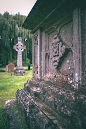 An ornate stone monument with intricate carvings stands prominently in the foreground, surrounded by lush green grass. In the background, a Celtic cross headstone is visible next to a large weeping willow tree.