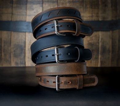A stack of leather belts in various shades of brown and black, each with visible stitching and silver buckles, arranged against a wooden background.
