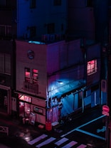 A building on a street corner is illuminated by neon pink and blue lights, creating a cyberpunk aesthetic. The reflections on the wet ground add to the futuristic atmosphere. The building has signs in a foreign language, an entrance visible on the ground floor, and a few windows with glowing lights. Bicycles are parked against the wall.