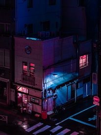 A building on a street corner is illuminated by neon pink and blue lights, creating a cyberpunk aesthetic. The reflections on the wet ground add to the futuristic atmosphere. The building has signs in a foreign language, an entrance visible on the ground floor, and a few windows with glowing lights. Bicycles are parked against the wall.