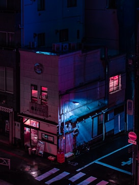 A building on a street corner is illuminated by neon pink and blue lights, creating a cyberpunk aesthetic. The reflections on the wet ground add to the futuristic atmosphere. The building has signs in a foreign language, an entrance visible on the ground floor, and a few windows with glowing lights. Bicycles are parked against the wall.