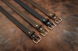 Four leather belts arranged in parallel, each featuring silver metal buckles. The belts are placed on a textured brown leather surface, showcasing different shades of brown and black.