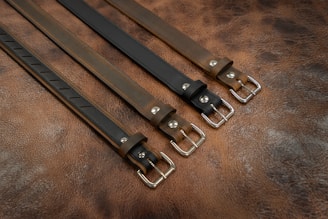 Four leather belts arranged in parallel, each featuring silver metal buckles. The belts are placed on a textured brown leather surface, showcasing different shades of brown and black.