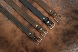 Four leather belts are laid out parallel to each other on a textured brown leather surface. Each belt features a metallic buckle and a different shade of brown or black, with a smooth, polished finish.