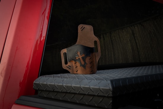 A leather holster with a cross design, featuring a mix of brown and black leather, is placed on top of a diamond-patterned metal surface. The background includes a red part of a vehicle and a dark reflective glass.