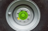 A close-up view of a large vehicle wheel with a prominently visible hub. The hub is painted bright green and is surrounded by a light grey metallic rim. Several lug nuts are visible, securing the wheel to the hub. The tire, which is black, is partially visible with intricate patterns and lettering embossed on its surface.