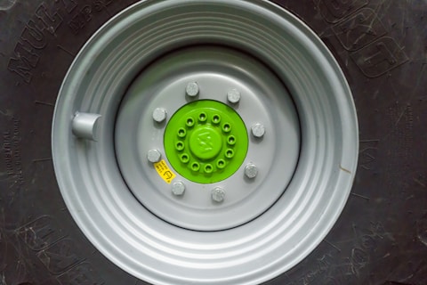 A close-up view of a large vehicle wheel with a prominently visible hub. The hub is painted bright green and is surrounded by a light grey metallic rim. Several lug nuts are visible, securing the wheel to the hub. The tire, which is black, is partially visible with intricate patterns and lettering embossed on its surface.