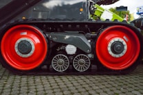 A close-up view of the track and wheel mechanism of a large track vehicle on a cobblestone surface. The track features two large, red wheels with metallic hubs and smaller, intricate metal components between them. The background shows parts of the vehicle with industrial elements.