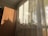 A wooden wardrobe stands next to a large window covered by sheer curtains. Sunlight filters through the window, casting interesting shadows and highlighting a variety of potted plants lined up on the windowsill. The setting suggests a cozy, sunlit room.