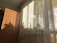 A wooden wardrobe stands next to a large window covered by sheer curtains. Sunlight filters through the window, casting interesting shadows and highlighting a variety of potted plants lined up on the windowsill. The setting suggests a cozy, sunlit room.
