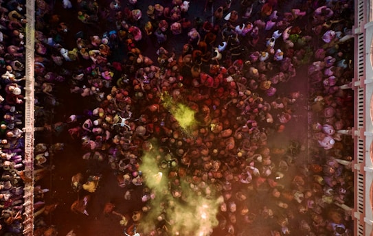 A vibrant overhead view of a densely packed crowd of people engaged in a lively celebration, with colorful powders dispersed in the air, possibly indicating a festival like Holi. The scene is bustling with energy as people gather in an urban setting between two buildings.