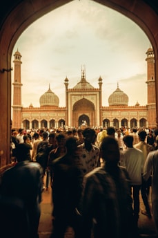An ornate mosque stands prominently with tall minarets and large domes under a cloudy sky. A large crowd of people, some blurred in motion, move through an arched gateway towards the mosque.