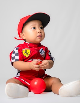 A baby is wearing a red racing-themed outfit with a red cap. The outfit features colorful patches, including a checkered design and logo. The child is sitting on the floor and holding a small red ball, gazing upwards with a curious expression. The background is a plain white.