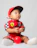 A baby is wearing a red racing-themed outfit with a red cap. The outfit features colorful patches, including a checkered design and logo. The child is sitting on the floor and holding a small red ball, gazing upwards with a curious expression. The background is a plain white.