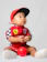 A baby is wearing a red racing-themed outfit with a red cap. The outfit features colorful patches, including a checkered design and logo. The child is sitting on the floor and holding a small red ball, gazing upwards with a curious expression. The background is a plain white.