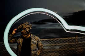 A person wearing camouflage clothing and a tactical helmet with night vision goggles stands in front of a chain-link fence. The background is a dark coastal landscape with blurred waves. A long exposure effect creates a glowing curved light trail above the person.