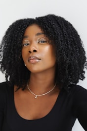 A woman looking confidently at the camera with curly black hair and glossy skin. She wears shimmering eye makeup and a small, delicate necklace. The background is a plain white, highlighting her features.