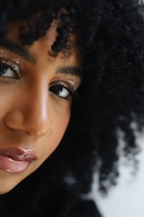 A close-up of an individual with curly hair, featuring glittery eye makeup, a nose ring, and glossy lips. The lighting emphasizes the texture of the hair and the shimmer of the makeup.