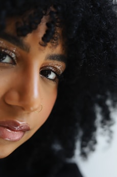 A close-up of an individual with curly hair, featuring glittery eye makeup, a nose ring, and glossy lips. The lighting emphasizes the texture of the hair and the shimmer of the makeup.