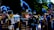 A marching band in blue and gold uniforms is performing. The members are playing various musical instruments such as flutes, trumpets, and drums. They are wearing elaborate hats with large blue plumes.