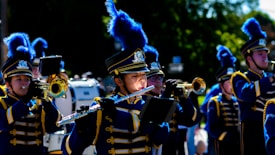 A marching band in blue and gold uniforms is performing. The members are playing various musical instruments such as flutes, trumpets, and drums. They are wearing elaborate hats with large blue plumes.