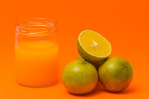 A clear glass jar filled with orange juice is positioned to the left. Next to it, three whole green oranges are stacked, with one cut in half, revealing its juicy, pulp-filled interior.