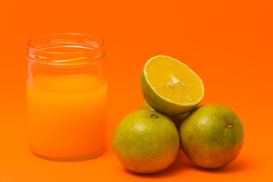 A clear glass jar filled with orange juice is positioned to the left. Next to it, three whole green oranges are stacked, with one cut in half, revealing its juicy, pulp-filled interior.