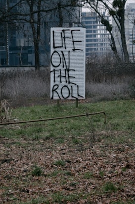 A sign with text 'LIFE ON THE ROLL' painted over what appears to be a weathered, text-filled billboard standing in a desolate field. The surrounding area is overgrown with grass and fallen leaves, and bare trees are visible in the background, with urban buildings in the distant background adding to the scene's neglected feel.