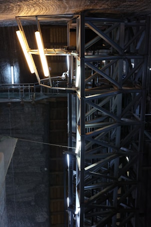 A large industrial structure with metal beams and bright fluorescent lighting arranged vertically. The setting appears to be an indoor environment with dim lighting and partially visible walls. The metallic beams form a framework that supports the lighting, which casts a stark white glow against the dark surroundings.