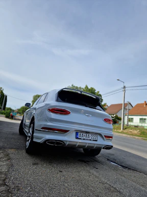 A white luxury SUV with a sleek design is parked on the side of a road. The vehicle has a European license plate and is surrounded by a suburban environment, including houses and greenery. Overhead, the sky is clear, creating a calm atmosphere.