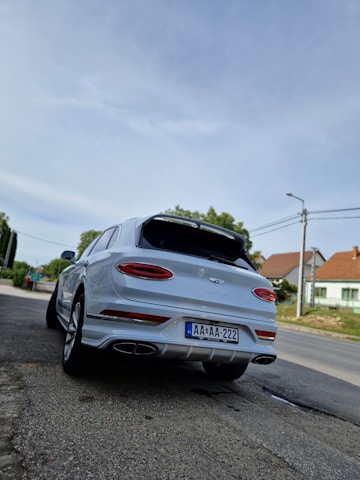 A white luxury SUV with a sleek design is parked on the side of a road. The vehicle has a European license plate and is surrounded by a suburban environment, including houses and greenery. Overhead, the sky is clear, creating a calm atmosphere.