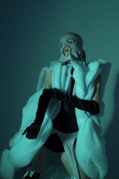 A person is wearing a balaclava mask and a luxurious, oversized white fur coat, complemented by black gloves and a black and white outfit underneath. The lighting casts a greenish hue, adding a mysterious and avant-garde atmosphere.