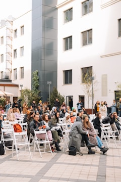 A gathering of people seated on white folding chairs in an outdoor urban setting. The audience is made up of individuals dressed in casual and cozy attire, including coats and scarves, suggesting cool weather. The surrounding area features modern building architecture with large windows, minimalistic decoration, and some greenery placed around.