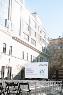 An outdoor event setup with rows of white and black chairs arranged in front of a stage. A large sign on the stage reads 'Festival of creative industries' with a date '09.09'. The venue is surrounded by modern architecture with large windows and a brick building visible in the background. Plants and trees add a natural touch to the urban setting.