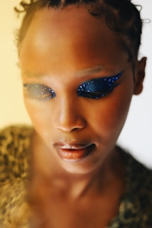 A close-up portrait of a person looking downward with intricate blue eye makeup adorned with small jewels. The makeup accentuates the eyes with a bold design. The overall lighting is warm and soft, creating a gentle and serene atmosphere.