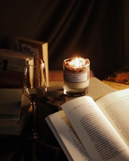 A cozy setting with a lit candle, an open book, and a wine glass. Several books are stacked next to them, creating a warm and inviting atmosphere.