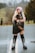A person with long, colorful hair stands on a wet road holding a fluffy cat. They are wearing a black outfit and distinctive knee-high boots with metal buckles. The background features an out-of-focus rural setting with trees and grass.