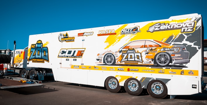 A large white trailer with vibrant graphics featuring a stylized car and various logos. The graphics include the text 'NZ Zoo Performance' and 'Central Drift Team'. The trailer is parked on a sunny day with a clear blue sky, showcasing its detailed design and multiple wheels.
