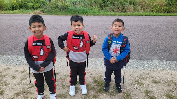 Three young children stand together outdoors on a gravel path with greenery and a paved road in the background. They are smiling and wearing backpacks with superhero-themed designs, featuring Spider-Man and Captain America. The children are dressed in casual outfits suitable for play or a school day.