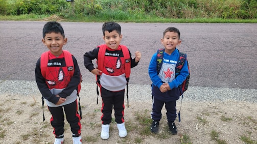 Three young children stand together outdoors on a gravel path with greenery and a paved road in the background. They are smiling and wearing backpacks with superhero-themed designs, featuring Spider-Man and Captain America. The children are dressed in casual outfits suitable for play or a school day.