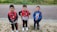 Three young children stand together outdoors on a gravel path with greenery and a paved road in the background. They are smiling and wearing backpacks with superhero-themed designs, featuring Spider-Man and Captain America. The children are dressed in casual outfits suitable for play or a school day.