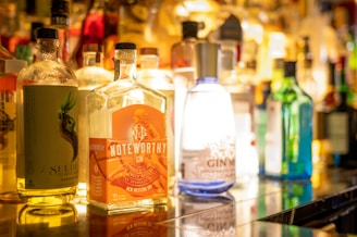 A variety of liquor bottles are arranged on a shelf, with prominent labels such as 'Noteworthy' gin. The focus is on a collection of colorful bottles, each uniquely designed with creative branding. The background is softly blurred, enhancing the vibrant colors and reflections.