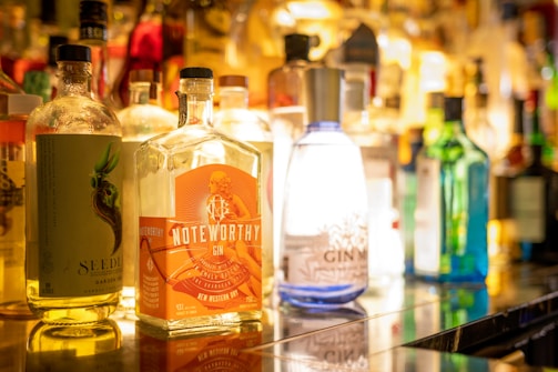 A variety of liquor bottles are arranged on a shelf, with prominent labels such as 'Noteworthy' gin. The focus is on a collection of colorful bottles, each uniquely designed with creative branding. The background is softly blurred, enhancing the vibrant colors and reflections.
