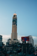 A tall skyscraper dominates the urban skyline with a large advertisement wrapped around its upper section. The surrounding area is bustling with dense city architecture, including older, lower-rise buildings. A prominent billboard for AirPods is visible in the foreground.