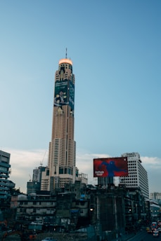 A tall skyscraper dominates the urban skyline with a large advertisement wrapped around its upper section. The surrounding area is bustling with dense city architecture, including older, lower-rise buildings. A prominent billboard for AirPods is visible in the foreground.