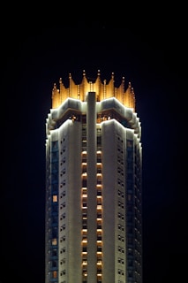 Tall building with a crown-like illuminated top, featuring bright lights and intricate architectural details. The structure stands out against a dark night sky.