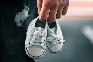 A hand holds a pair of small white baby shoes with laces. The background has a dark, textured surface with a torn area, adding contrast to the bright shoes.