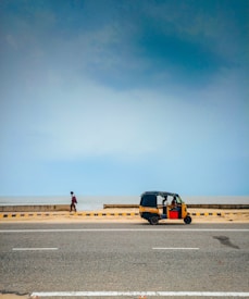 A coastal road with a person walking along the sidewalk next to a calm sea. A parked rickshaw is by the road, indicating a vibrant yet relaxed environment.