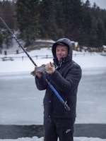 A person stands outdoors, holding a fishing rod and proudly displaying a fish in their hand. The surroundings are snowy, with a frozen body of water nearby and dense trees in the background. The person is dressed warmly in a black jacket with a hood.
